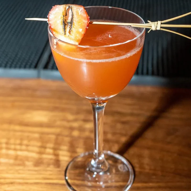 Orange-red cocktail in a coupe glass with grilled strawberry garnish.