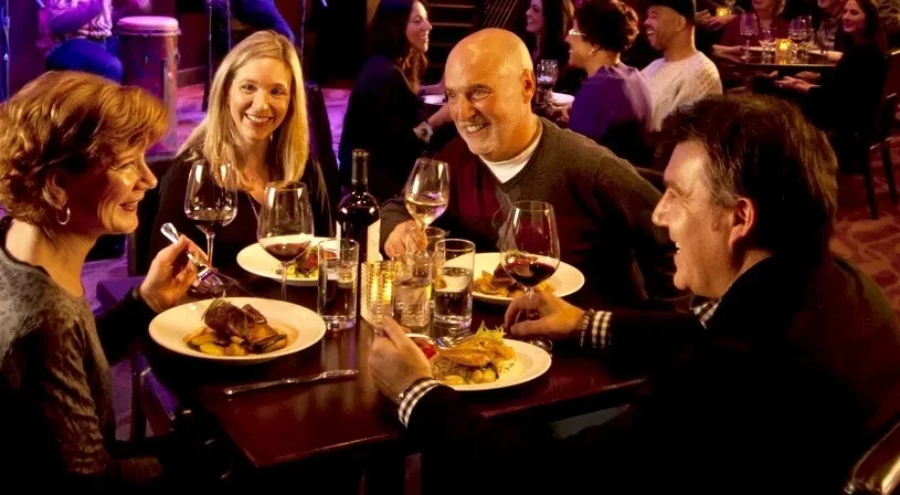 Four smiling people share a meal in a lively restaurant with a stage.