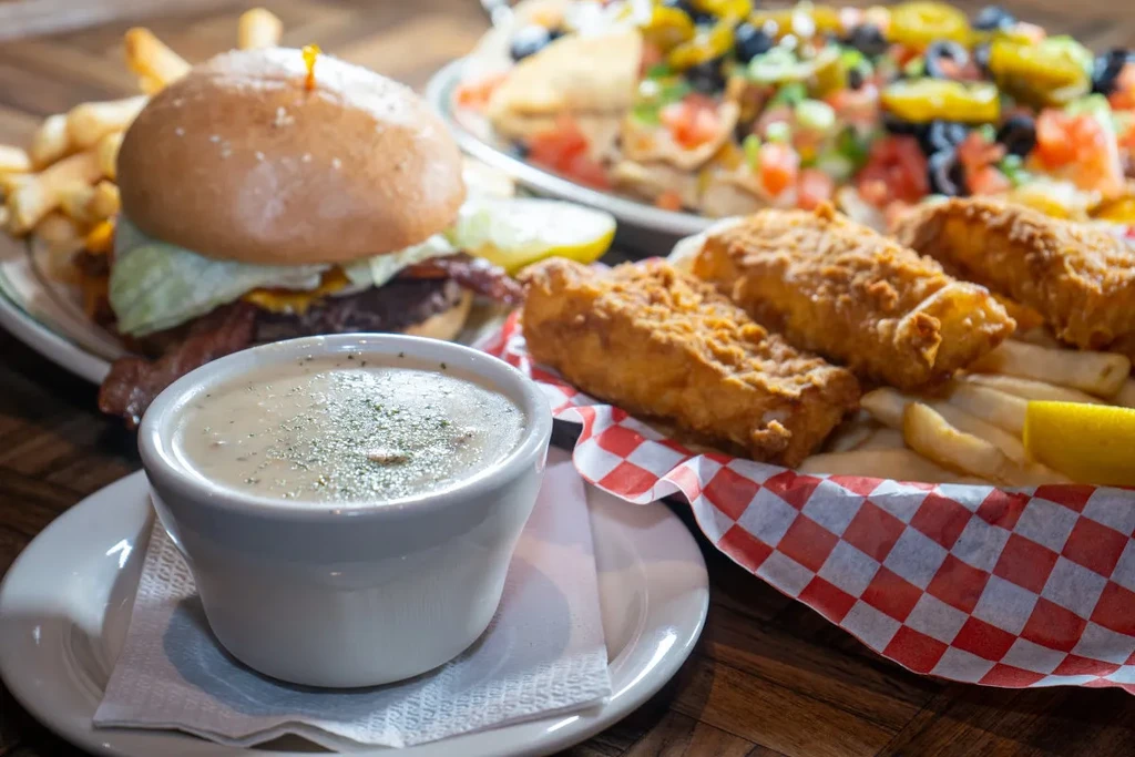 Clam chowder, fried fish, burger, and nachos on a wooden table.