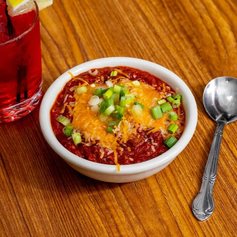 Bowl of chili with cheese, green onions, drink, and spoon on wooden table.