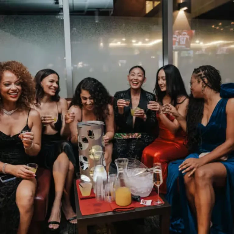 Six formally dressed women enjoying drinks in a festive bar.