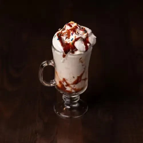 Creamy drink in glass mug, topped with whipped cream, syrup, sprinkles.