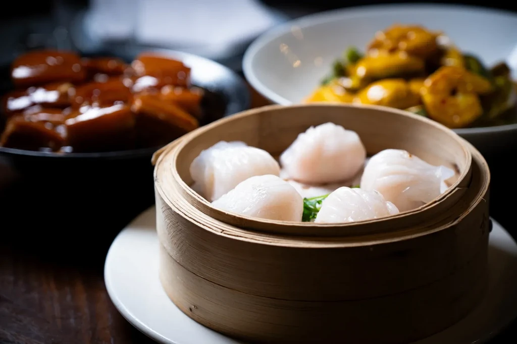 Six translucent shrimp dumplings in a steamer, with other dishes behind.