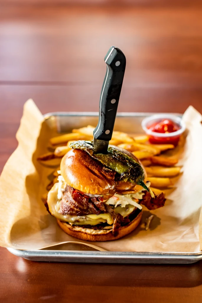 A knife-pierced burger with cheese, bacon, chili, and fries on a tray.