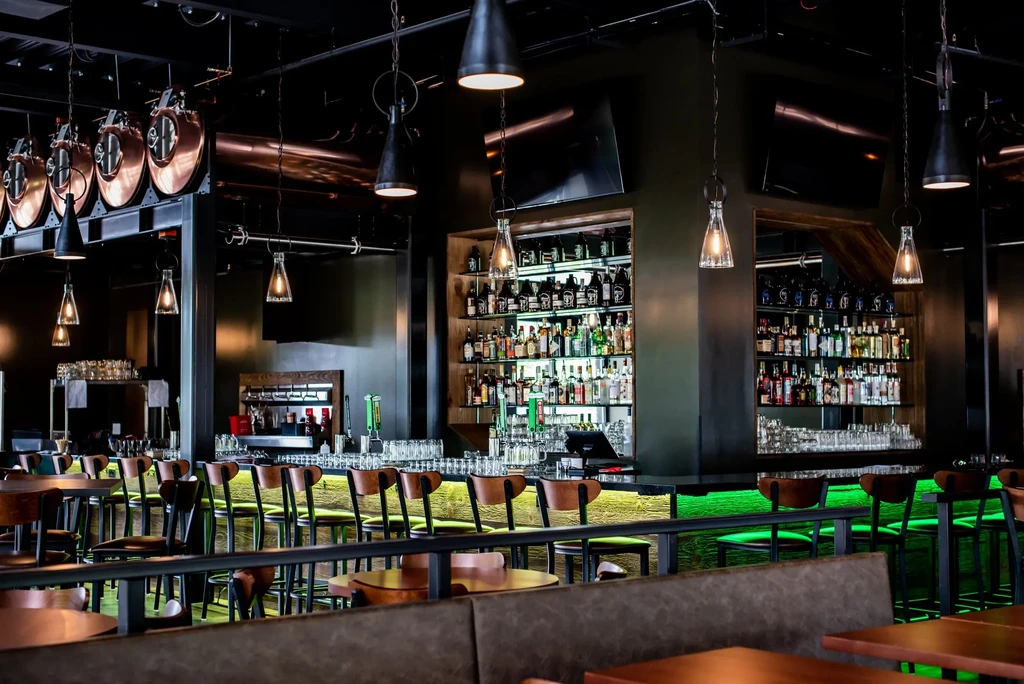 Modern bar with green underlit counter, liquor bottles, and industrial lighting.