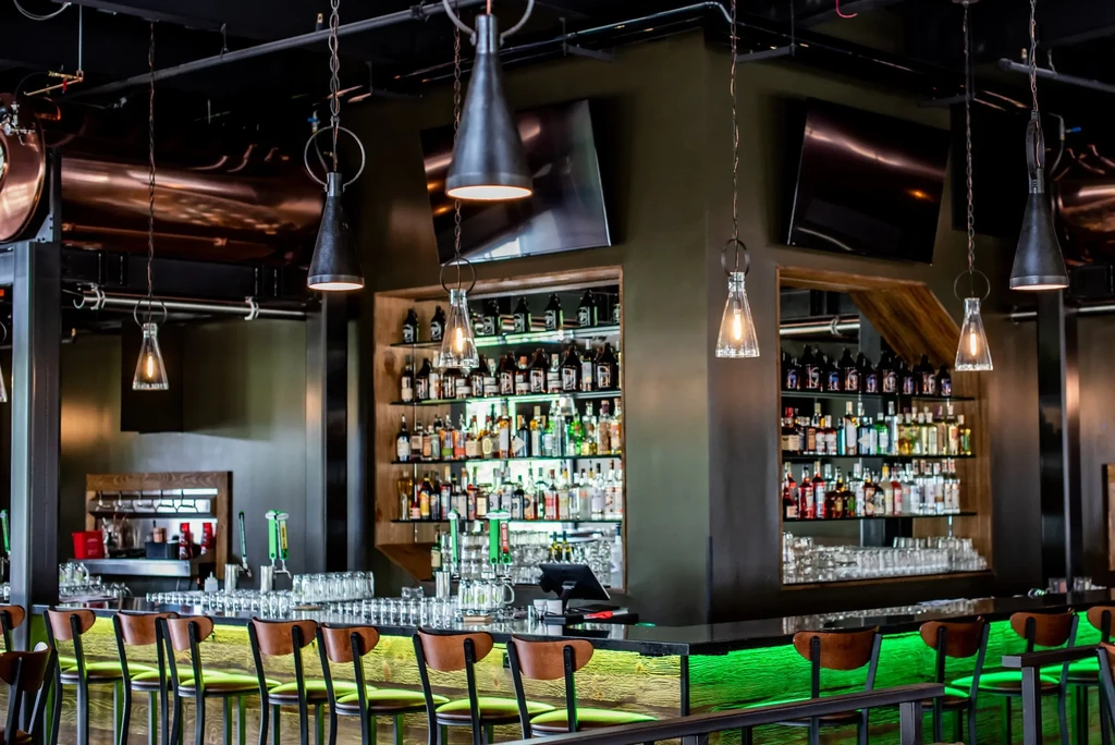 Modern bar with green-lit counter, stools, and extensive liquor bottles.