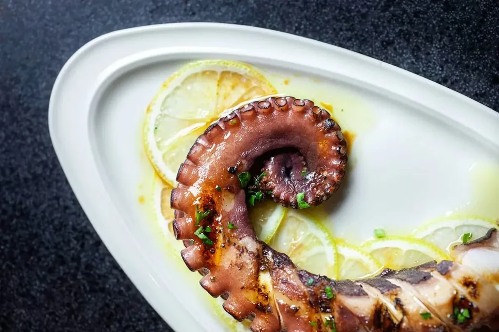 Charred grilled octopus tentacle, garnished with herbs and lemon slices.