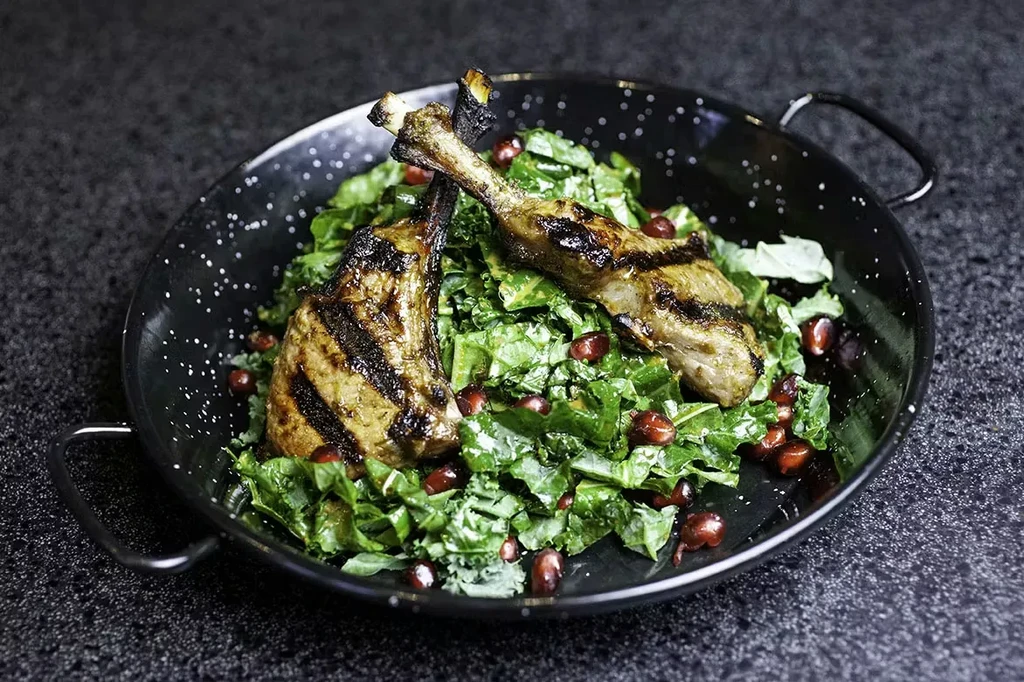 Grilled meat chops, greens, pomegranate seeds in dark skillet.