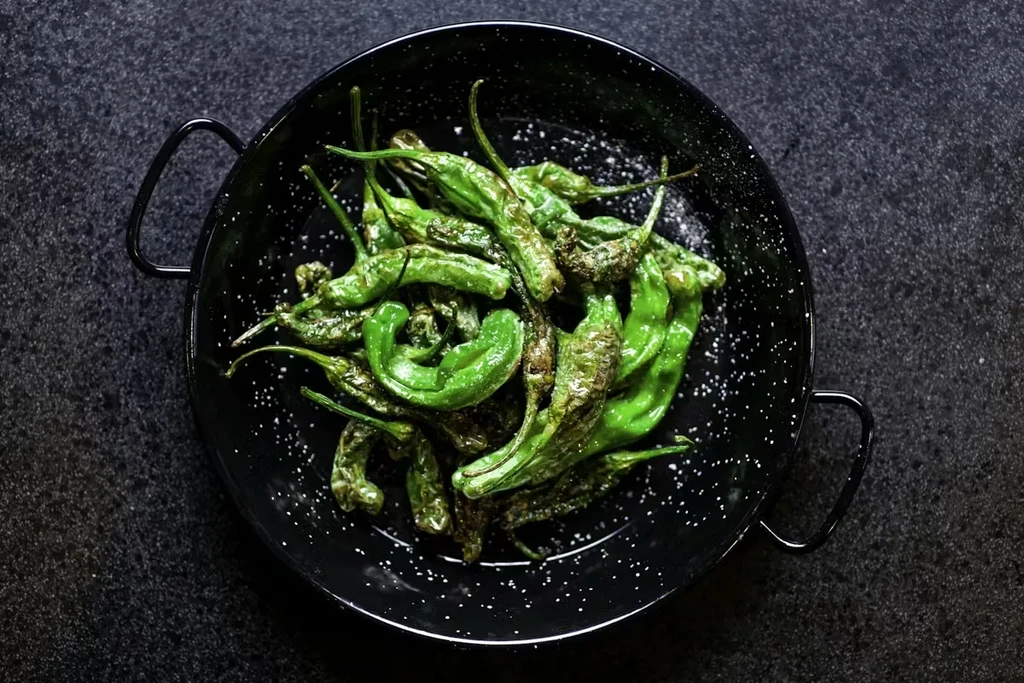 Blistered green peppers sprinkled with coarse salt in a black pan.