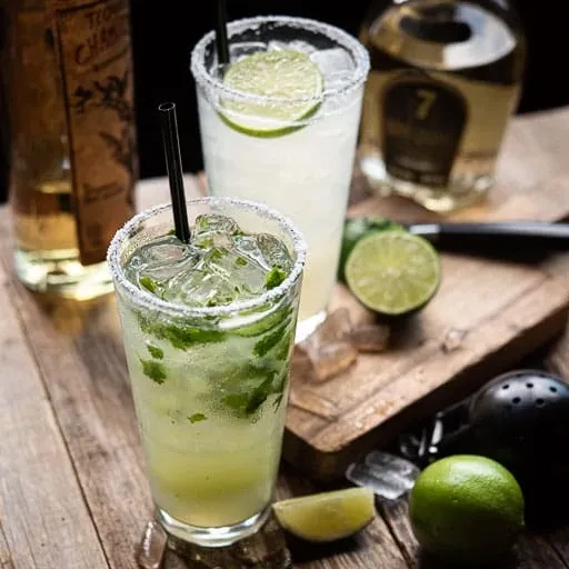 Two cocktails, limes, ice, and bar tools on a rustic wooden surface.