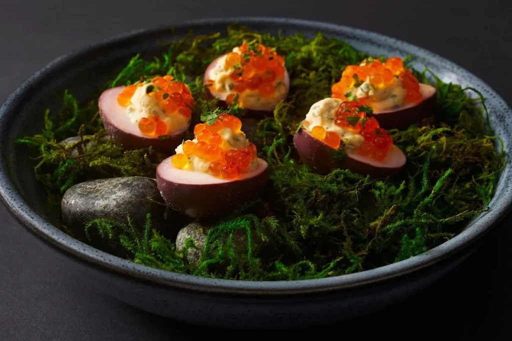 Five purple egg appetizers with orange roe on moss and stones.