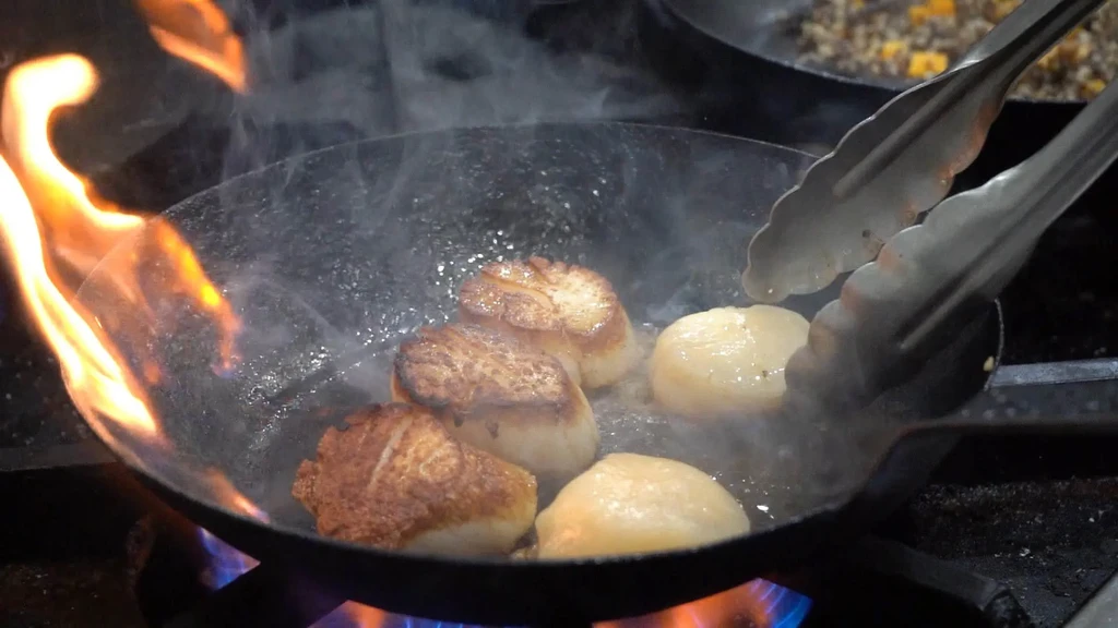 Scallops searing in a pan with dramatic orange flames and steam.