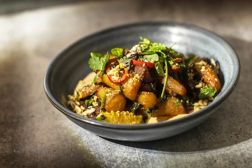 Roasted potato-like dish with herbs, chili, and peanuts in a dark bowl.