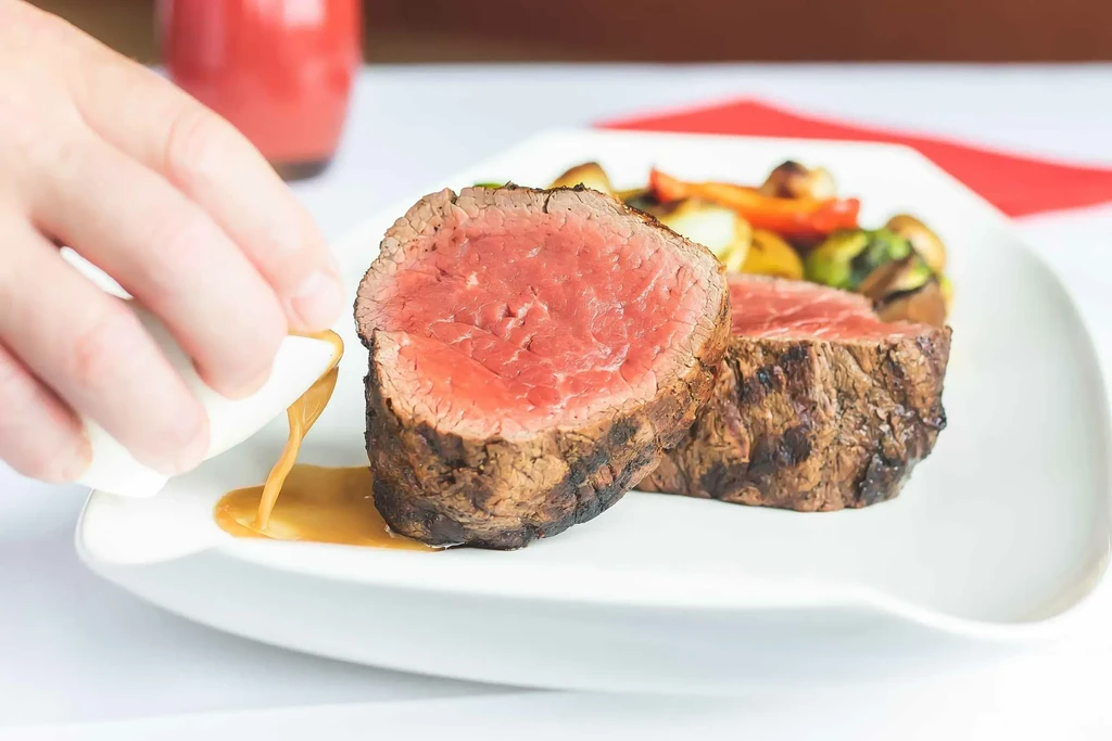 Hand pouring sauce over two medium-rare steaks with roasted vegetables.