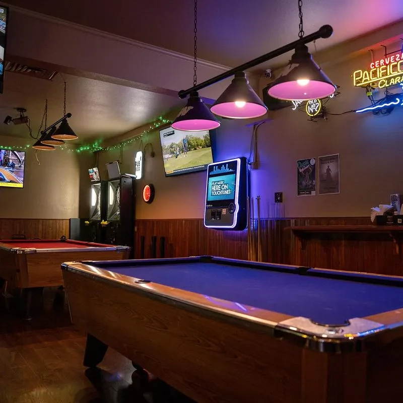 Two pool tables, purple lights, and neon signs in a dim bar.