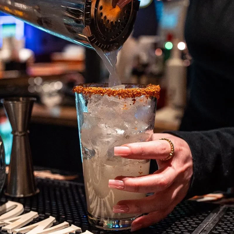 Hand with pink nails holds a red-rimmed drink, shaker pours liquid.