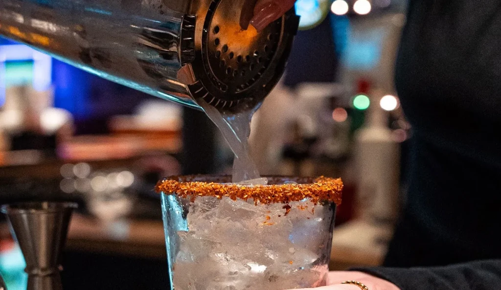 Cocktail shaker pouring drink into an ice-filled glass with a spiced rim.