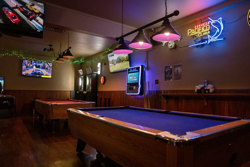 Two illuminated pool tables in a dim bar with sports on TVs.