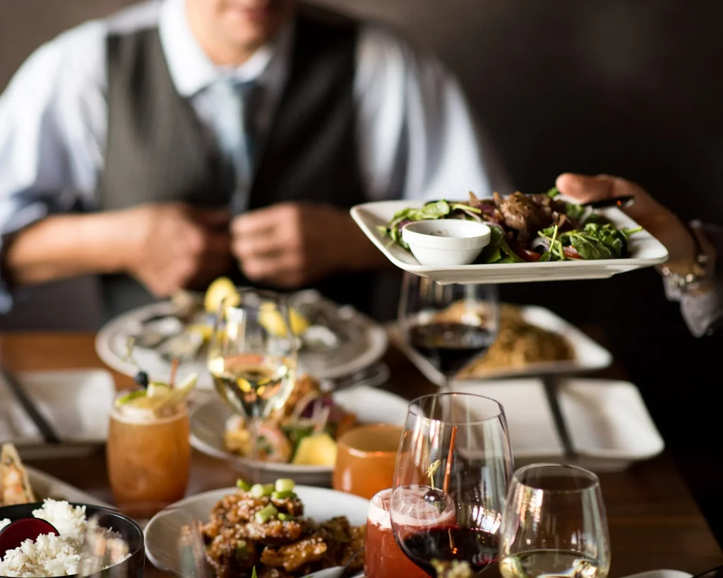 Server brings a salad to a table laden with food and drinks.