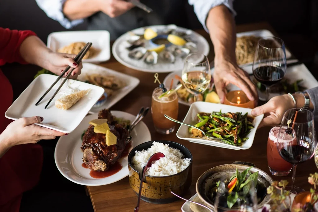 High-angle table shot: roasted meat, oysters, rice, drinks, and hands.