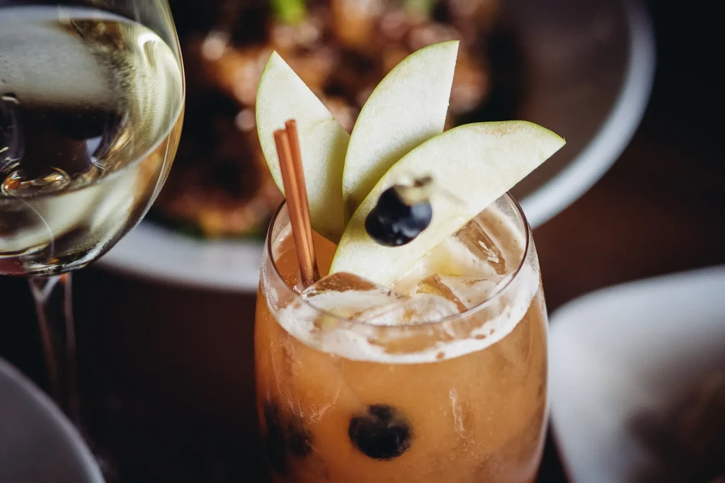 Tall amber cocktail with apple, cinnamon, berry, beside a white wine glass.