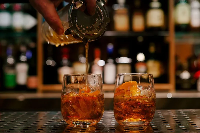 Amber cocktail poured into glass; another prepared nearby on a bar.
