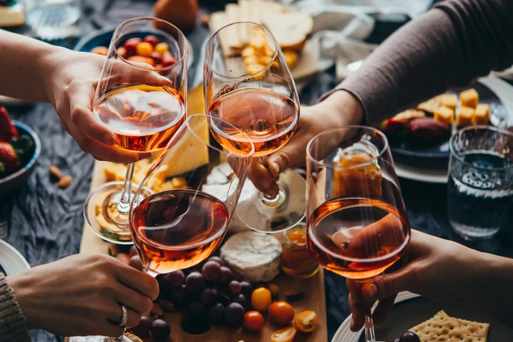Four hands clinking rosé wine glasses above a charcuterie spread.