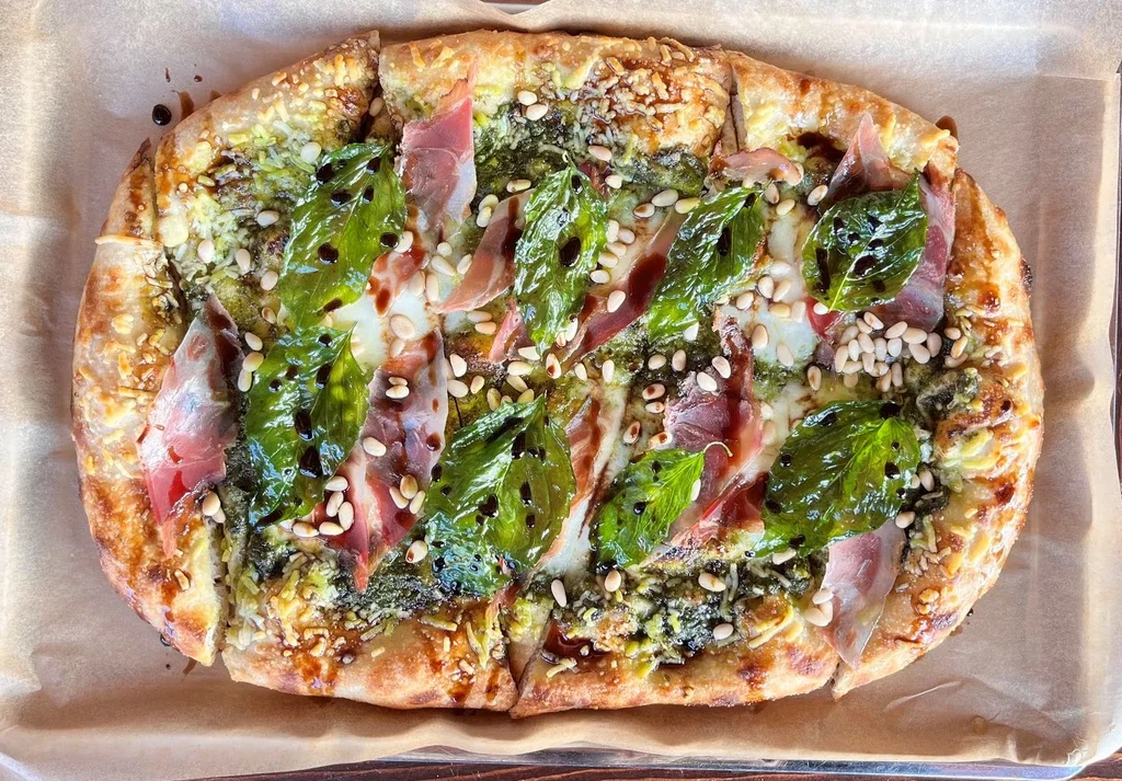Rectangular pesto pizza with prosciutto, mozzarella, basil, and pine nuts.