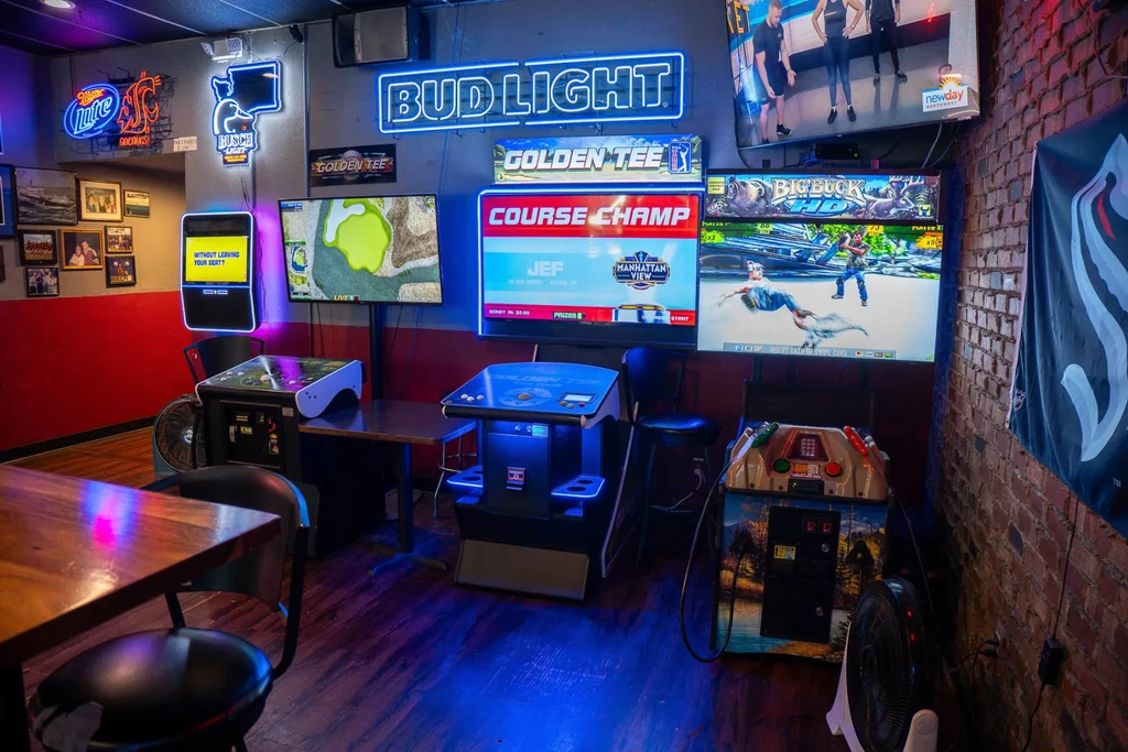 Dimly lit game room with arcades, TVs, and glowing neon beer signs.