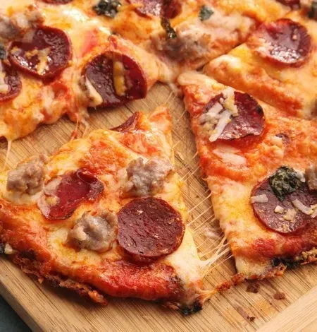 Close-up pizza: pepperoni, sausage, cheese pull, green herbs on wooden board.