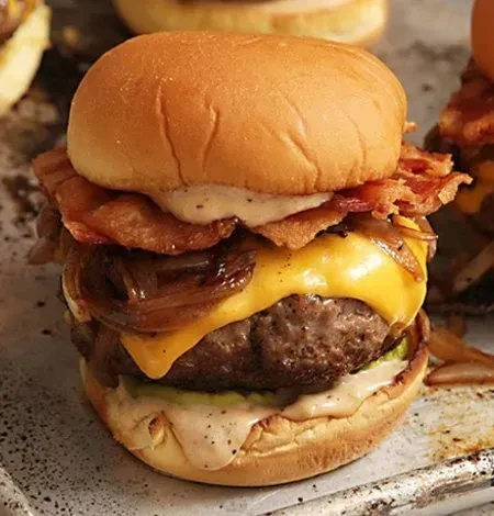 Gourmet burger close-up: beef, cheddar, bacon, caramelized onions, pickles, sauce.