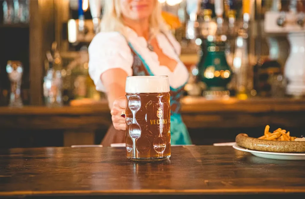 Person holding VELTINS beer stein, sausage, and fries on a bar.