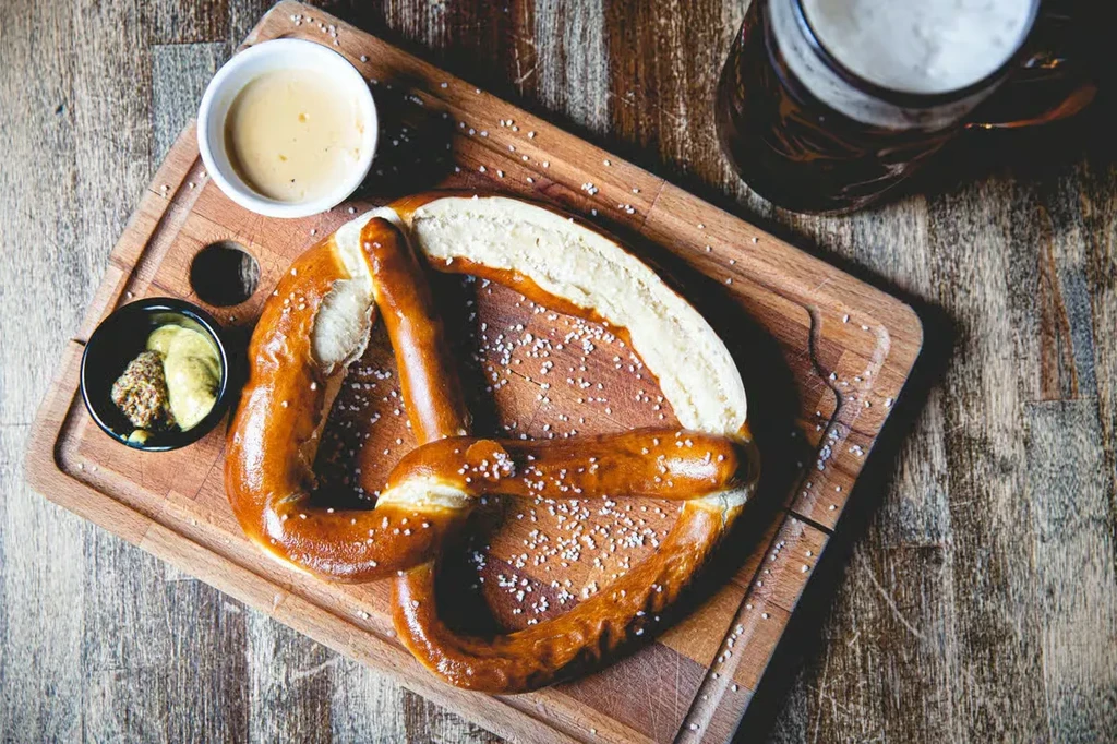 Top-down view: salted pretzel, two dips, and beer on wooden board.