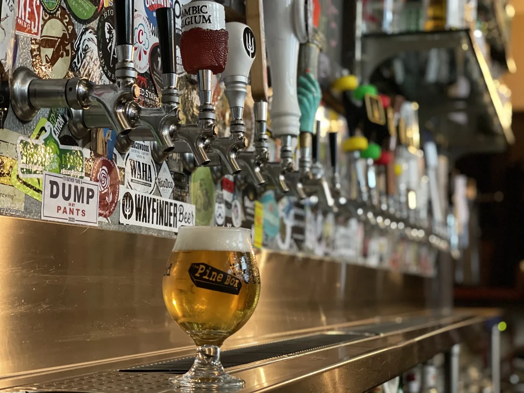 Glass of 'The Pine Box' beer, bar taps, and brewery stickers.