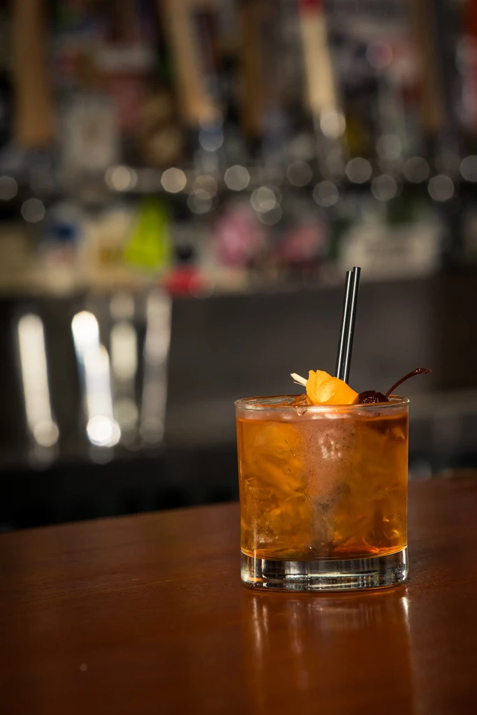 Garnished amber cocktail with ice, straw on dark wood bar.