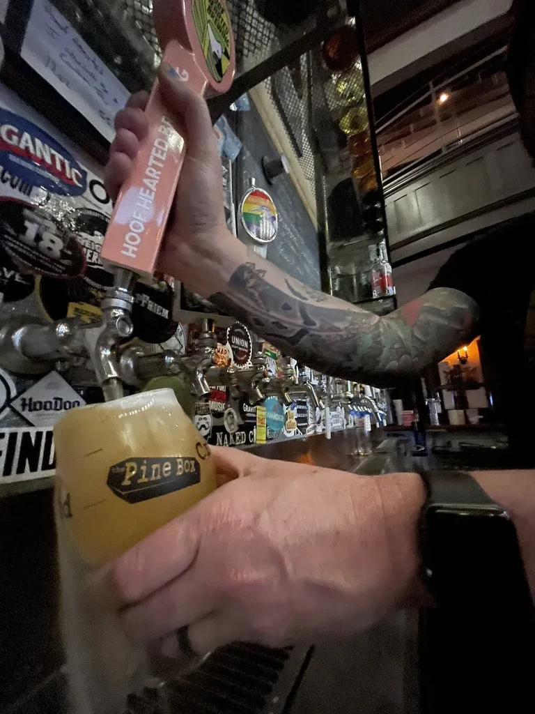 Tattooed person pours beer from tap into 'The Pine Box' glass.