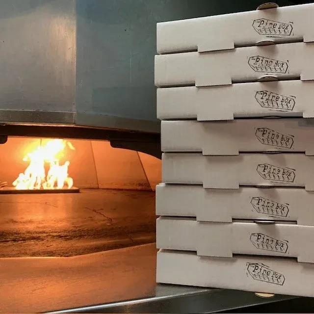 Industrial pizza oven with flames and stack of "Pine box" pizza boxes.