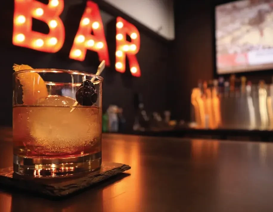 Amber cocktail with orange peel, dark cherry, and round ice at bar.