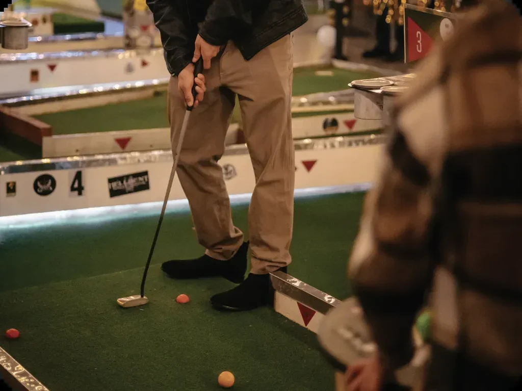 Person in tan pants putts orange golf ball on indoor mini-golf course.