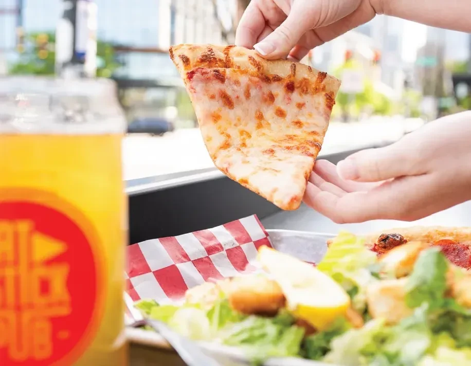 Hand holding pizza slice at 'BIG RIG PUB' with beer and salad.