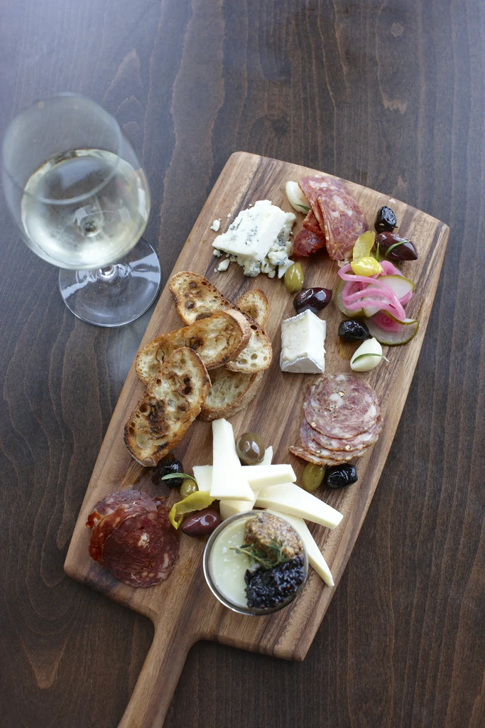 Wooden charcuterie board with cheeses, meats, bread, olives, and white wine.