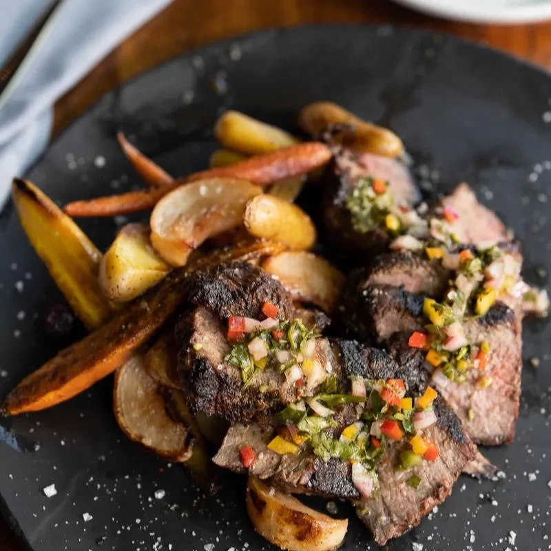 Plated grilled meat, green sauce, pepper relish, roasted potatoes, carrots.