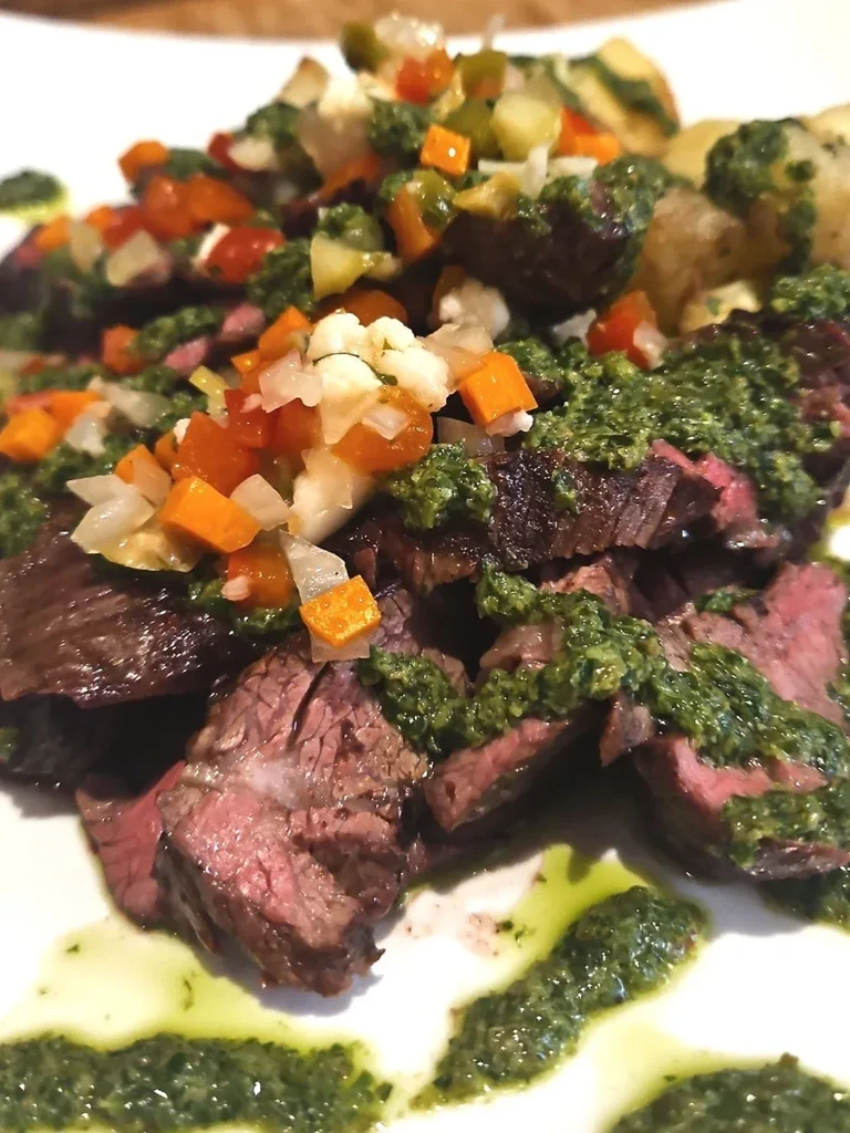 Sliced medium-rare steak with green chimichurri and diced vegetable relish.