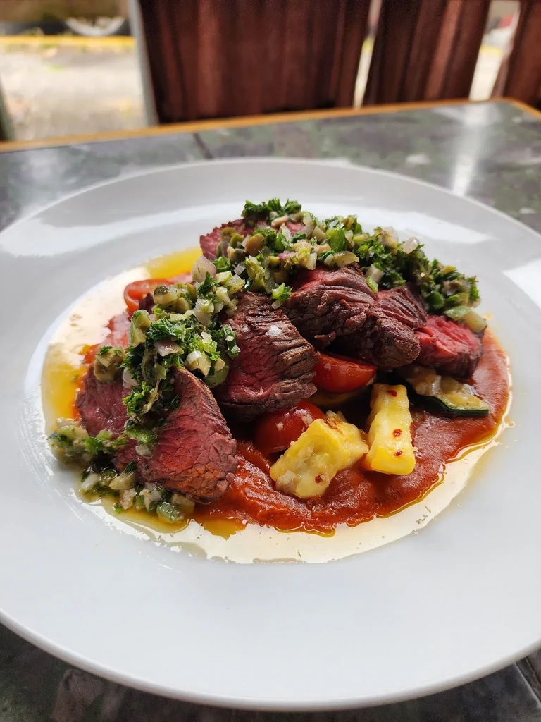 Plated medium-rare steak slices with green chimichurri, tomatoes, and vegetables.