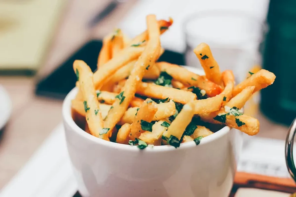 Close-up of golden french fries in a white bowl, garnished with green herbs.