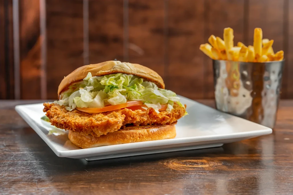 Large crispy fried chicken sandwich, lettuce, tomato, fries, on wood.