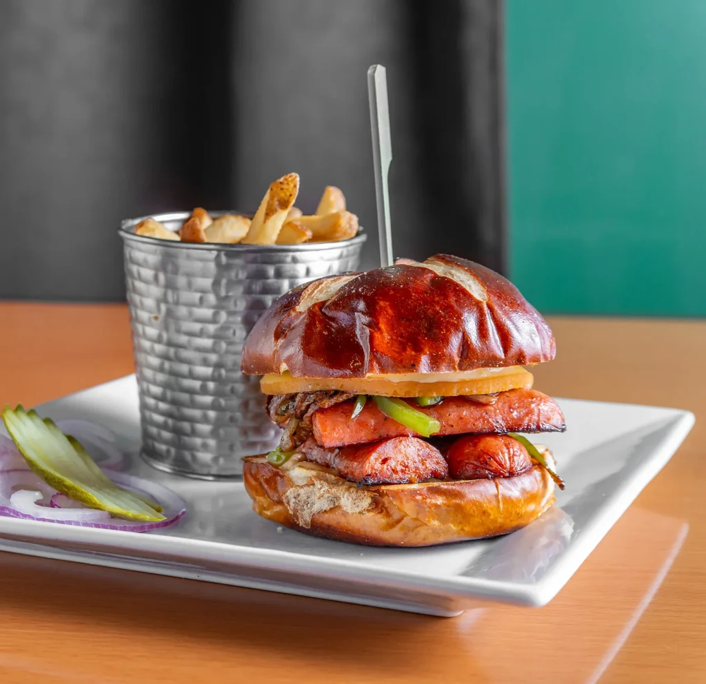 Pretzel bun sausage sandwich with caramelized onions, fries, and pickle.