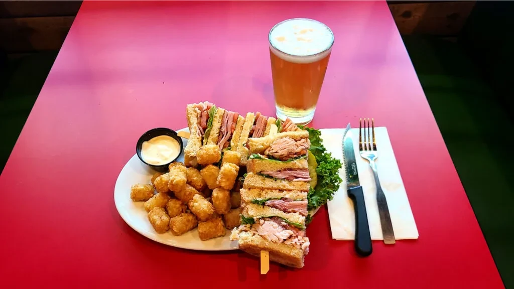 Overhead of a club sandwich, tater tots, and beer on red table.