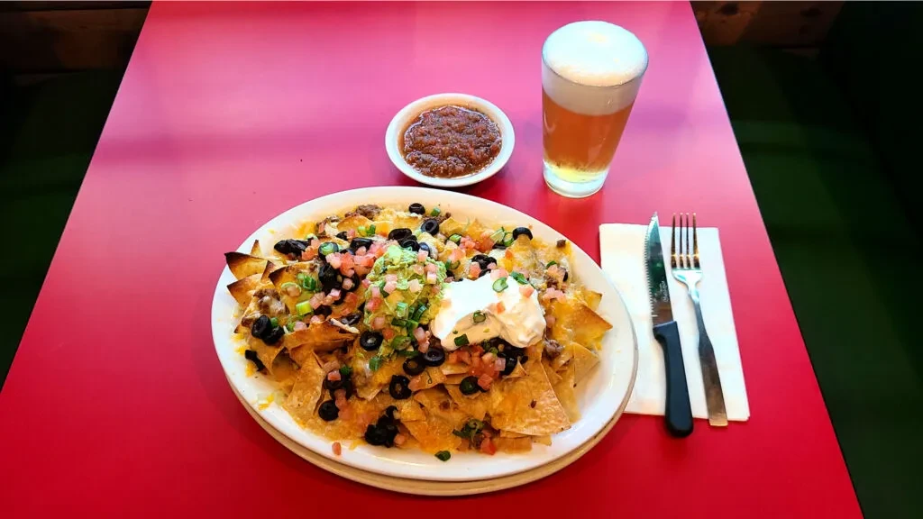 Overhead view of loaded nachos, salsa, and beer on red table.