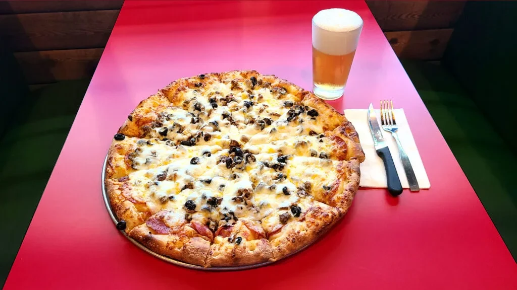 Multi-topping pizza, beer, and cutlery on a red table.